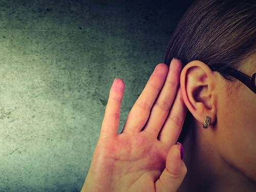 Woman holds her hand near ear and listens carefully isolated on gray wall background